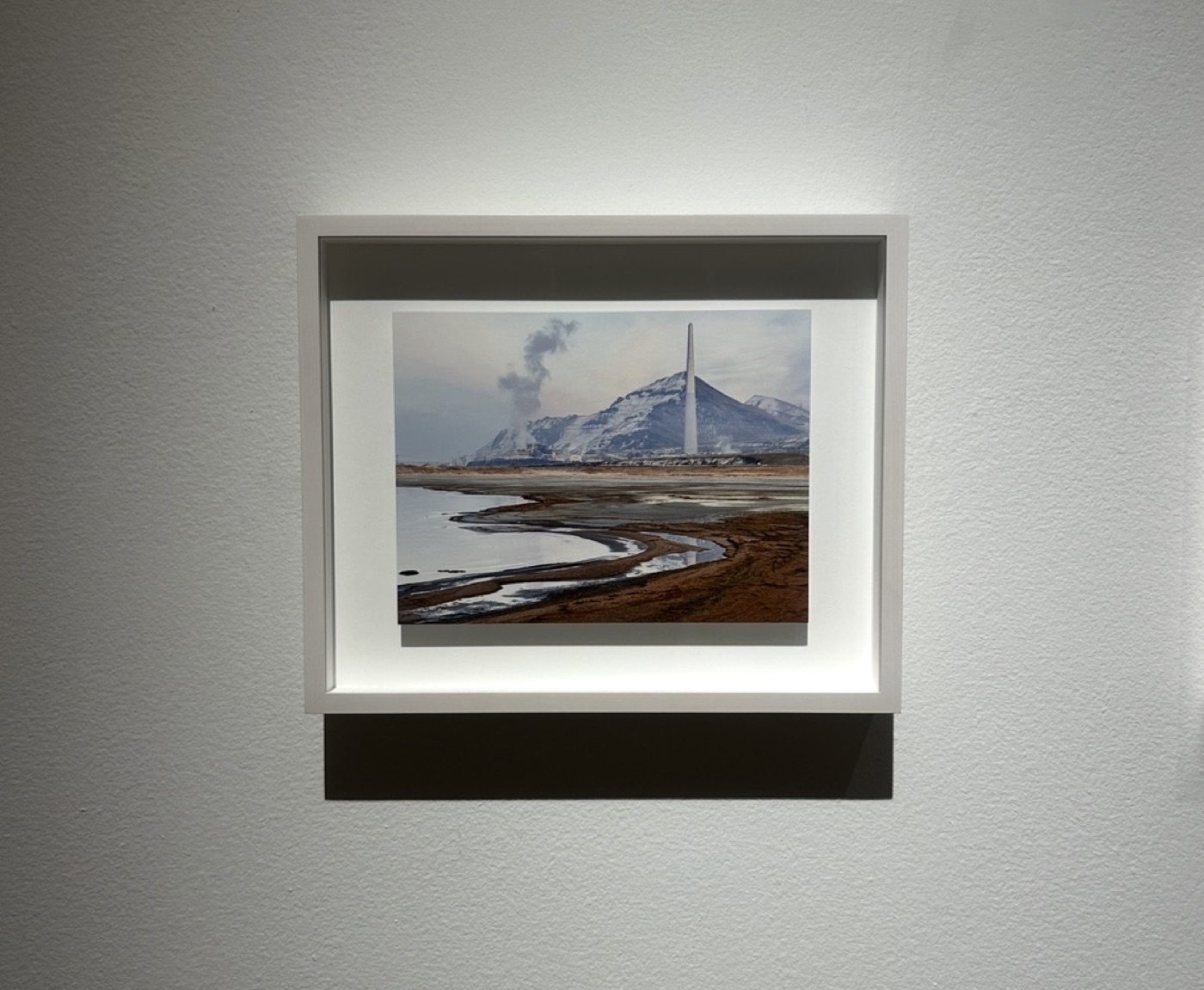 A framed photograph on a gallery wall: a smokestack and snowy mountain over a tidal shoreline