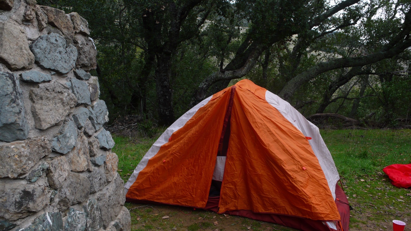 An orange tent pitched beside a stone chimney under oaks